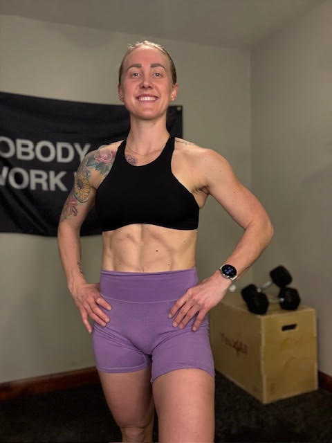Woman wearing a black sports bra and purple shorts, standing in a gym with dumbbells and a plyo box in the background.
